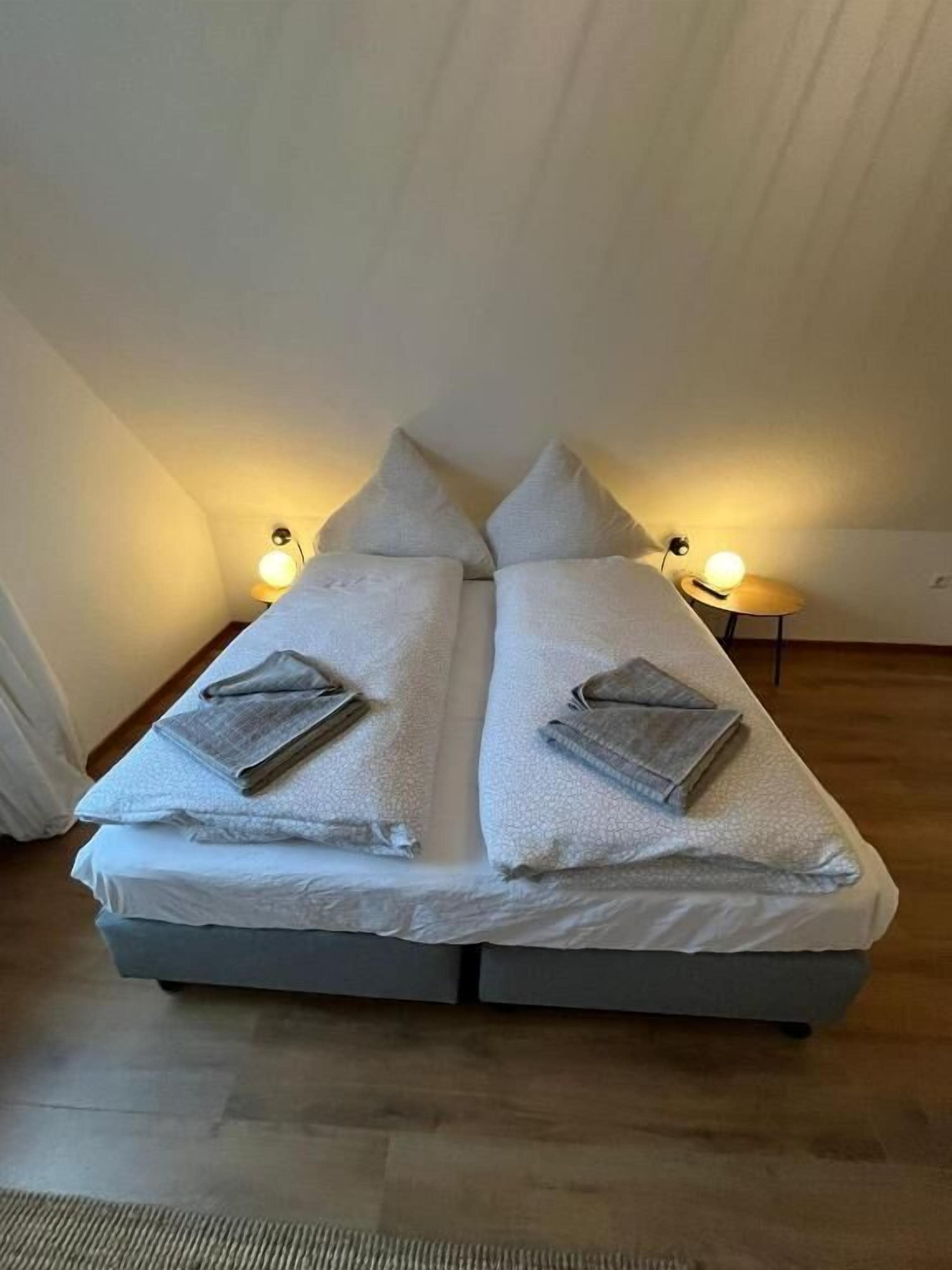 A twin bed with pillows and a blanket, two bedside lamps, and a wooden floor.