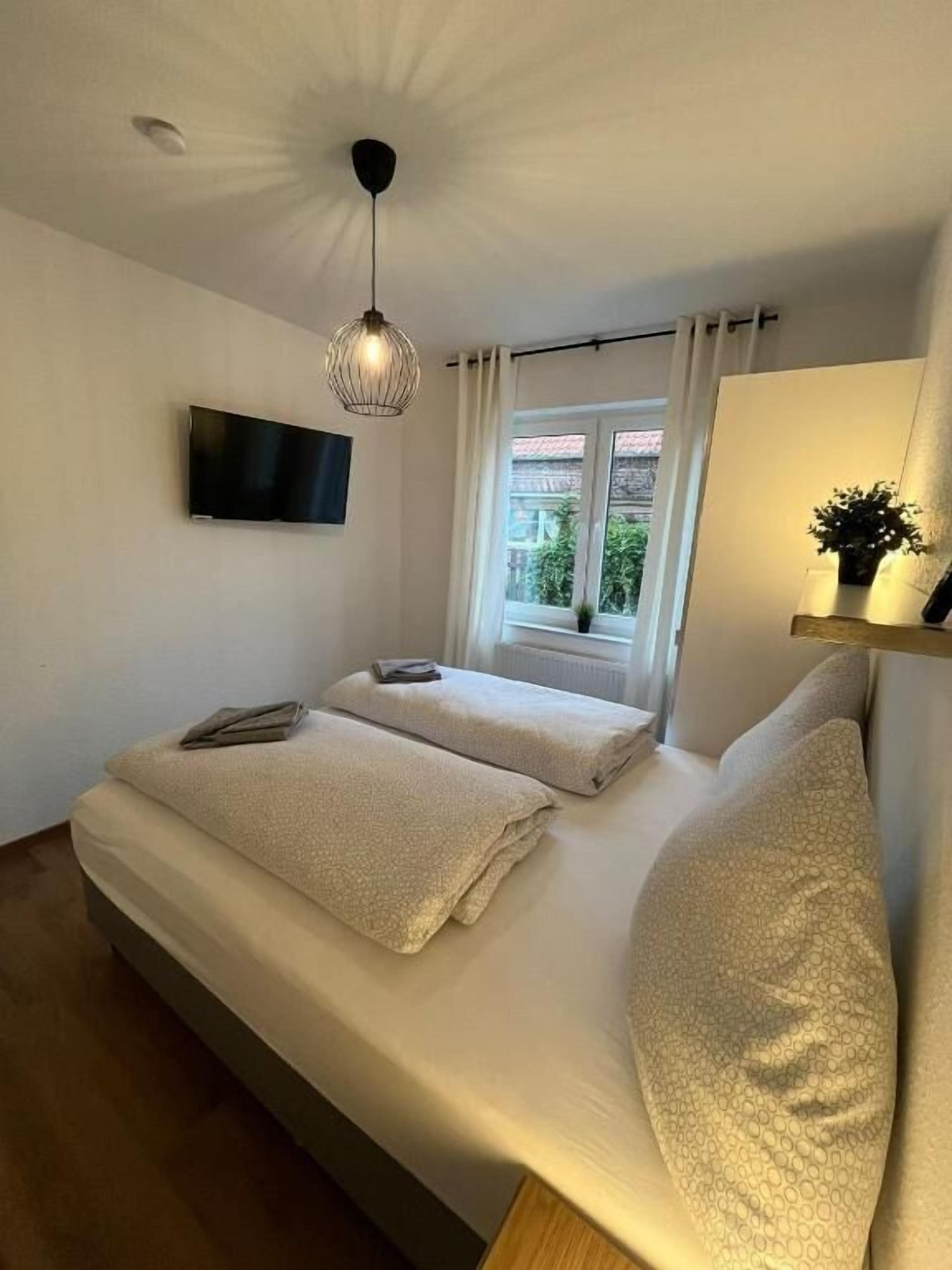 Ein Schlafzimmer mit einem Bett, einem an der Wand montierten Fernseher, einem Fenster mit Vorhängen und einer Deckenleuchte.