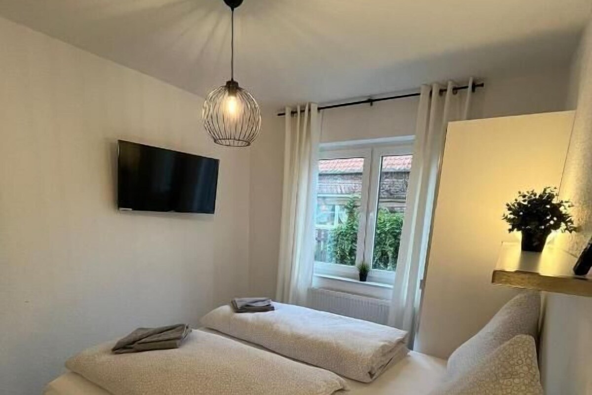 Une chambre à coucher avec un lit, un téléviseur fixé au mur, une fenêtre avec des rideaux et un luminaire au plafond.