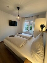 Ein Schlafzimmer mit einem Bett, einem an der Wand montierten Fernseher, einem Fenster mit Vorhängen und einer Deckenleuchte.