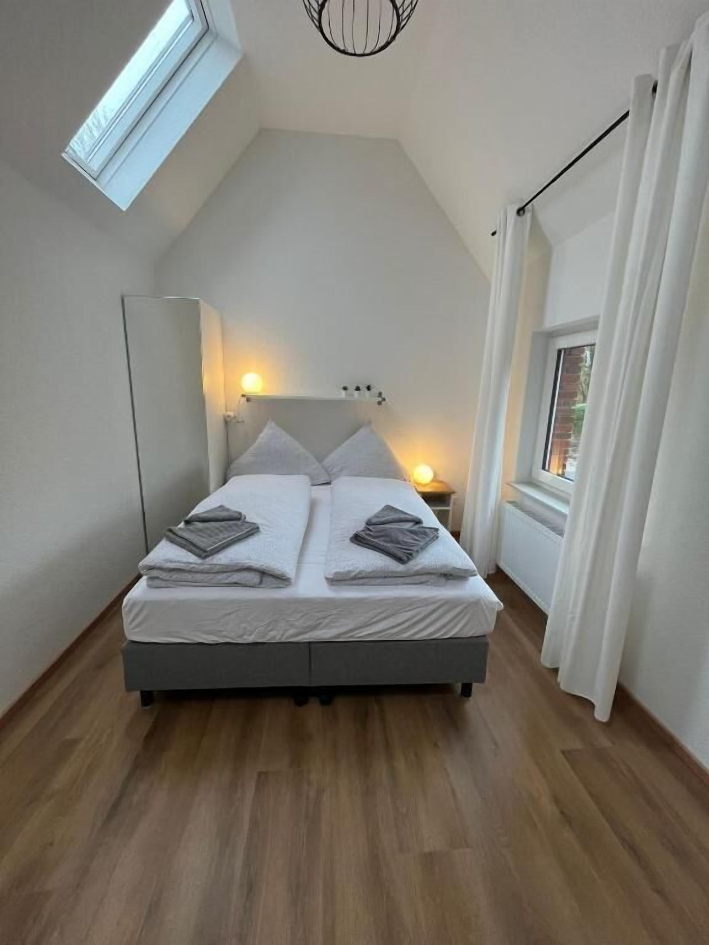Une chambre avec un plafond en pente, un lit avec des oreillers, une table de chevet avec une lampe et une fenêtre avec des rideaux.