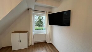 A room with a sloped ceiling, a white radiator, a white cabinet, a flat-screen TV mounted on the wall, and a window with curtains. - Ostfriesenperle (Aurich)