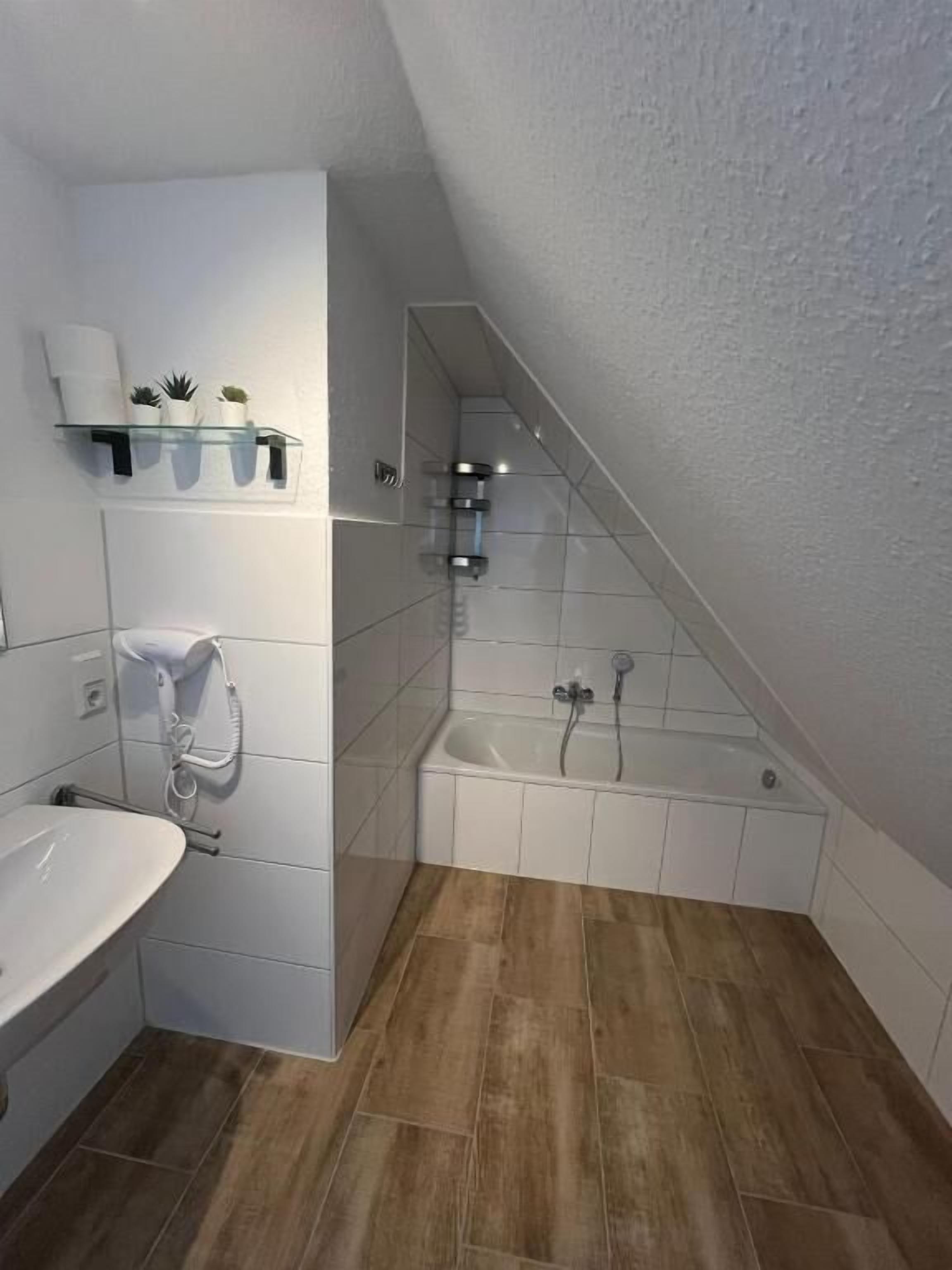 Ein Badezimmer mit einem weißen Waschbecken, einer Badewanne und einer Dusche. Es gibt Regale mit Pflanzen sowie einen Handtuchhalter.