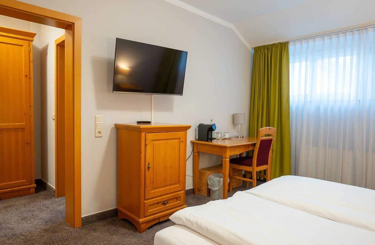 A hotel room with a bed, a desk with a chair, a television mounted on the wall, and a window with curtains.