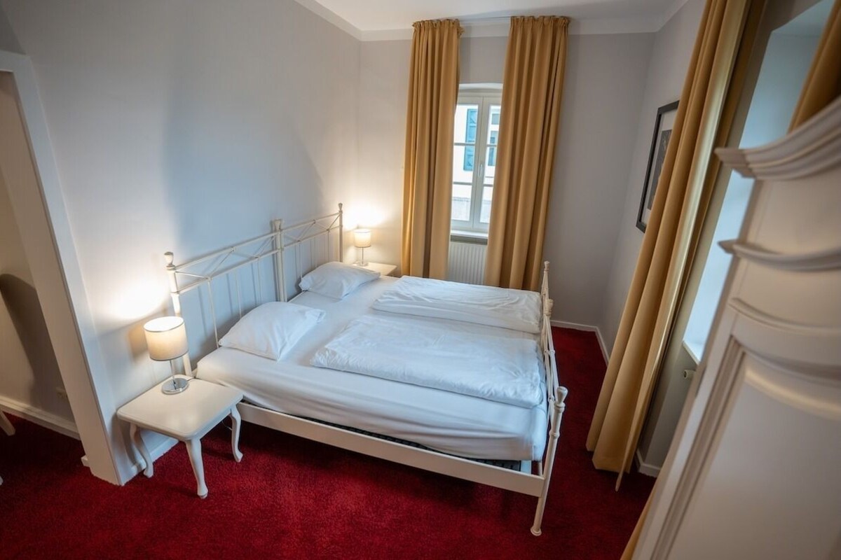 Una cama bien tendida con sábanas blancas, una mesita de noche con una lámpara y una ventana con cortinas.