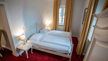 Ein ordentlich bezogenes Bett mit weißen Leinentüchern, ein Nachttisch mit einer Lampe und ein Fenster mit Vorhängen.