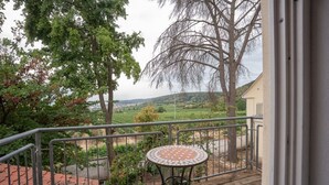 A balcony with a metal railing, a small round table, and a view of a vineyard and distant buildings.
