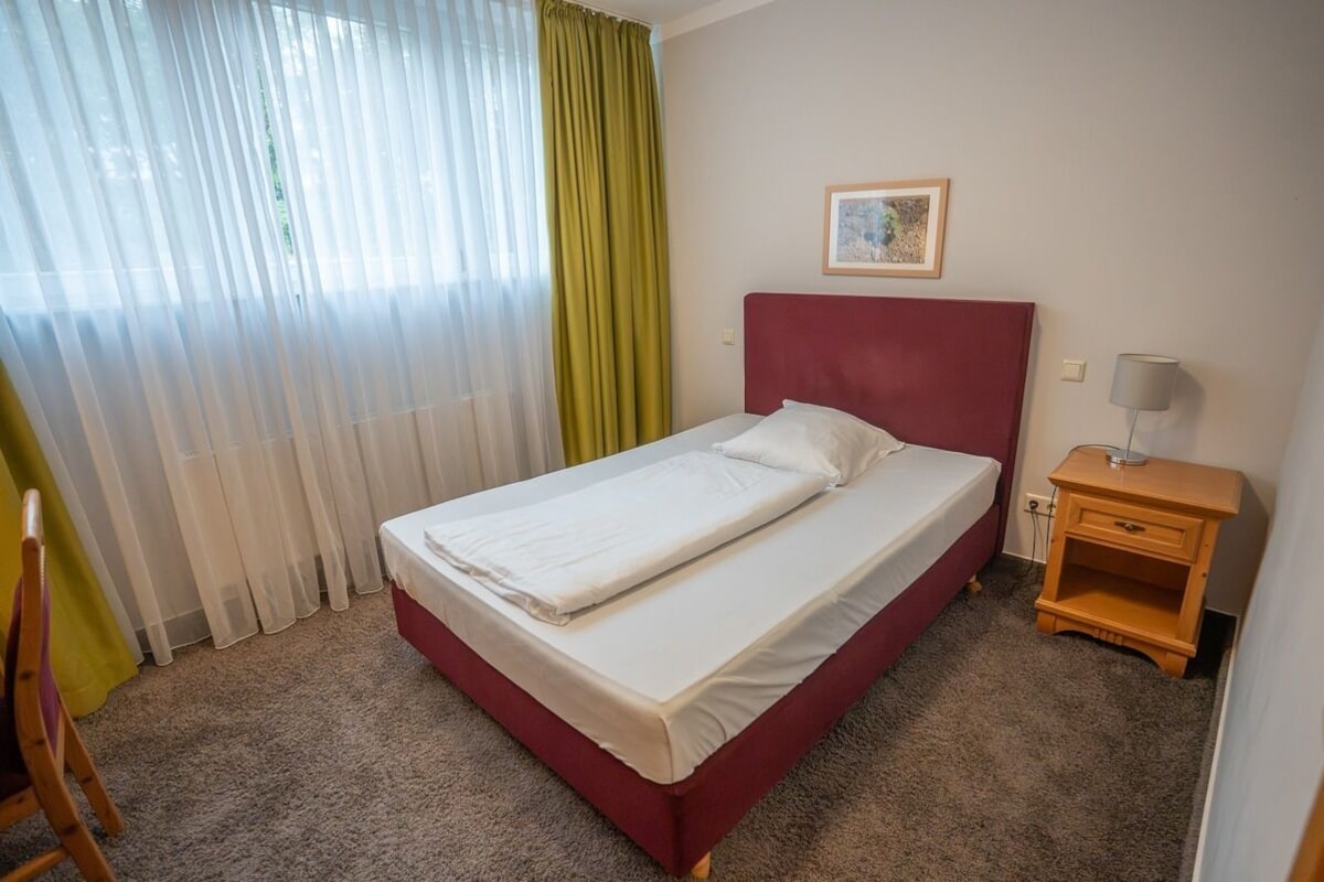 Una habitación de hotel con cama, mesita de noche, una silla y ventana con cortinas.