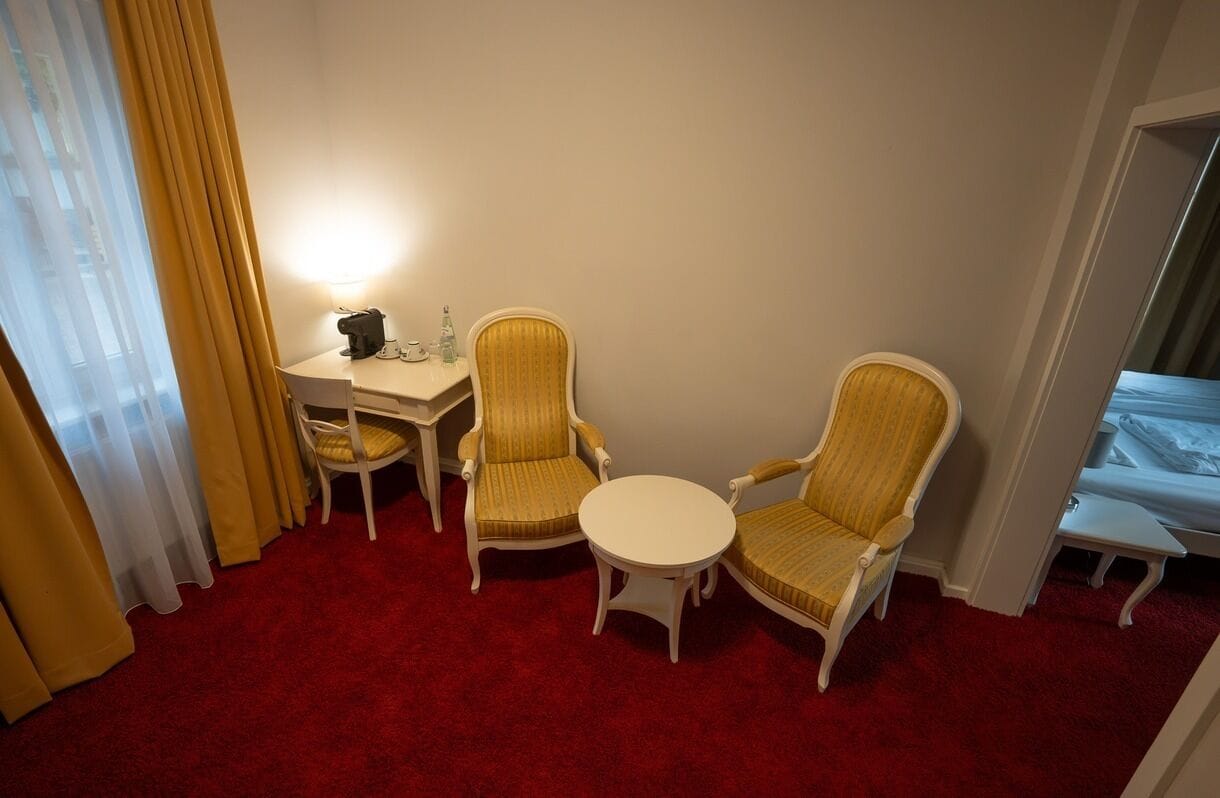 Ein Hotelzimmer mit rotem Teppich, zwei Sesseln, einem kleinen runden Tisch, einem Schreibtisch mit Lampe und einem Bett.