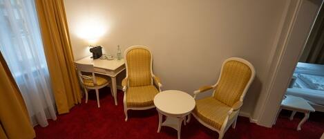 A hotel room with a red carpet, two armchairs, a small round table, a desk with a lamp, and a bed.