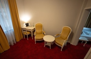 Habitación de hotel con alfombra roja, dos sillones, una mesita redonda, un escritorio con lámpara y una cama.