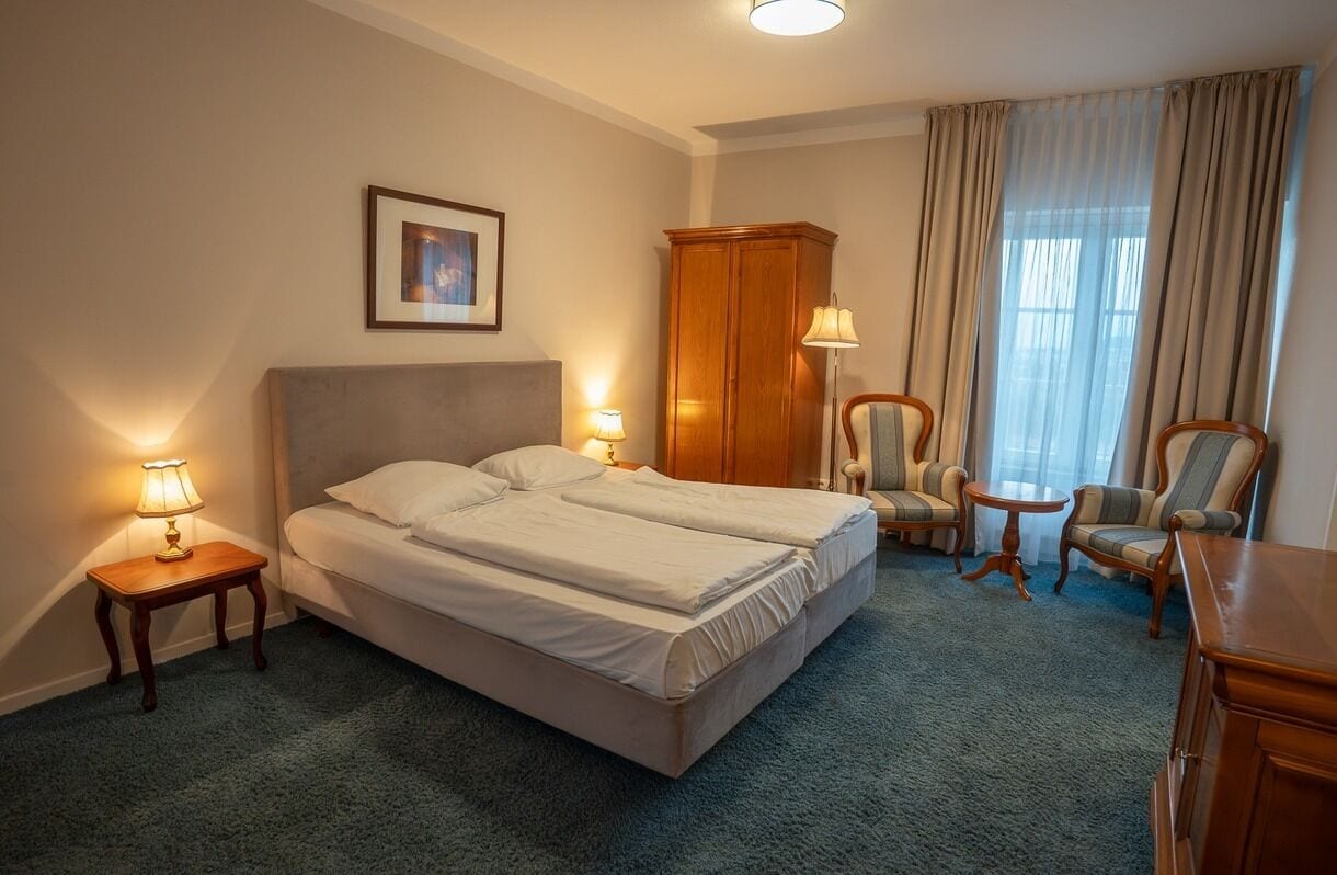 A hotel room with a double bed, two bedside lamps, a wooden wardrobe, a small round table, and two armchairs.