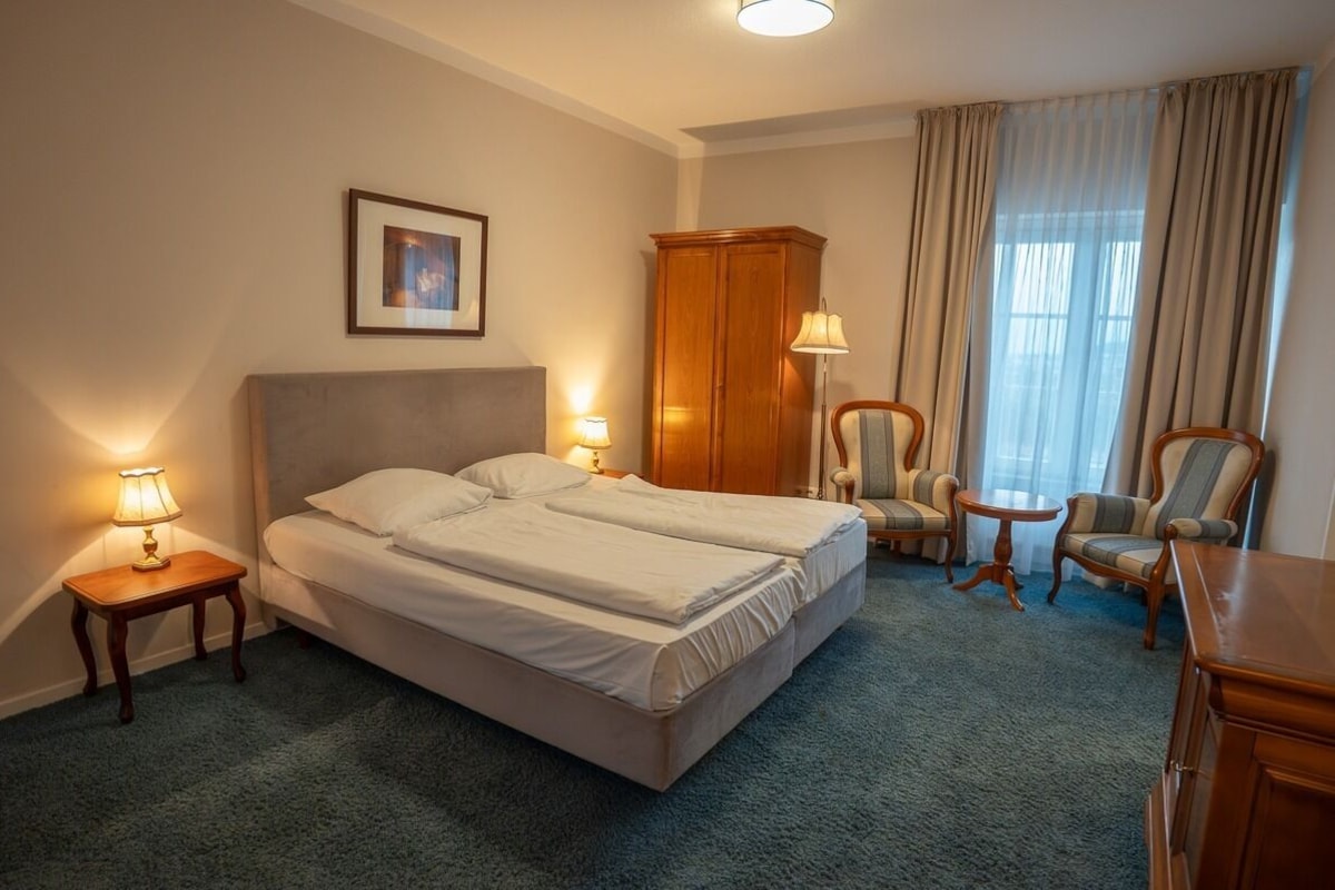 Habitación de hotel con cama doble, dos lámparas de noche, armario de madera, una mesita redonda y dos sillones.