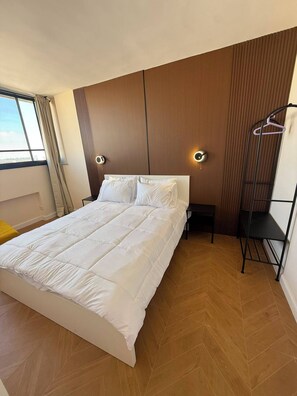 Une chambre moderne avec un grand lit, une table de chevet, une lampe fixée au mur et une fenêtre avec des rideaux.
