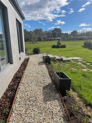 A modern building with large windows, a gravel pathway, and a grassy area with trees and shrubs.