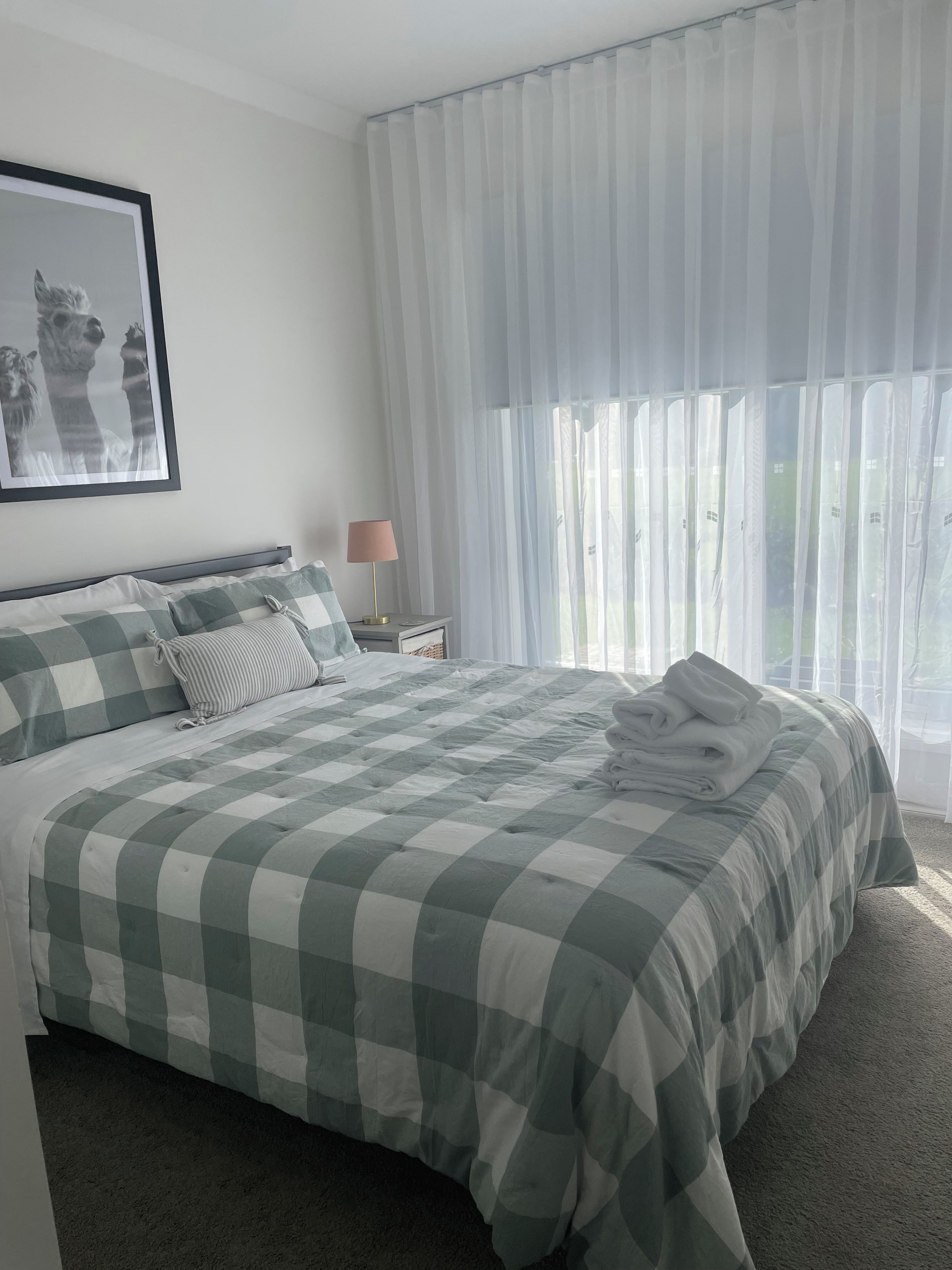 Quarto com cama grande, mesa de cabeceira com abajur, quadro na parede e cortinas leves.