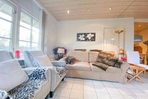 Ein modernes Wohnzimmer mit einer Couch, Sesseln und einem Couchtisch. Ein Fenster lässt natürliches Licht herein, an der Wand hängt ein Gemälde und auf einem Beistelltisch steht eine Lampe.