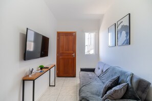 Ein modernes Wohnzimmer mit einem Flachbildfernseher, einer Holztür, einem kleinen Tisch mit einer Pflanze, einem grauen Sofa und zwei gerahmten Kunstwerken an der Wand.