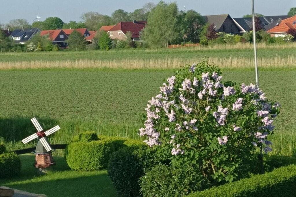 Een landelijk landschap met een windmolen, een bloeiende struik en huizen op de achtergrond.