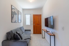 A living room with a grey sofa, a wooden door, a TV mounted on the wall, and a small table with a plant. - Comfortable Apartment 10min, Beach Jurere Jma009 (Florianópolis)