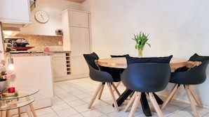 Uma sala de jantar com uma mesa redonda, quatro cadeiras, um vaso com plantas verdes e um relógio na parede.