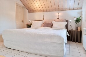 Un dormitorio con una cama grande, techo de madera, una puerta blanca y una mesita con un florero.