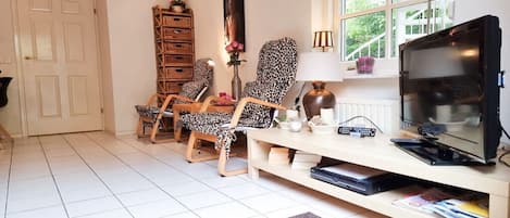 Een woonkamer met een televisie, twee fauteuils met luipaardprint, een houten kast en een raam met uitzicht op groen.