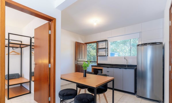 A compact kitchen with a wooden table, two black chairs, a black hanging plant, a stainless steel refrigerator, and a window with a view of trees. - Beautiful apt 10 min From the Beach, Jurere Jma010 (Florianópolis)