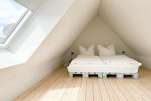 Un dormitorio con techo inclinado, tragaluz, cama con ropa blanca y suelo de madera.
