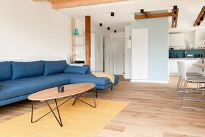 A modern living room with a blue sofa, a wooden coffee table, and a kitchen area in the background. - Holiday apartment with 3 bedrooms (Scharbeutz)