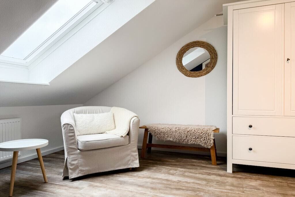 Una habitación con techo inclinado, un sofá blanco, un espejo redondo pequeño, un banco y un armario blanco.