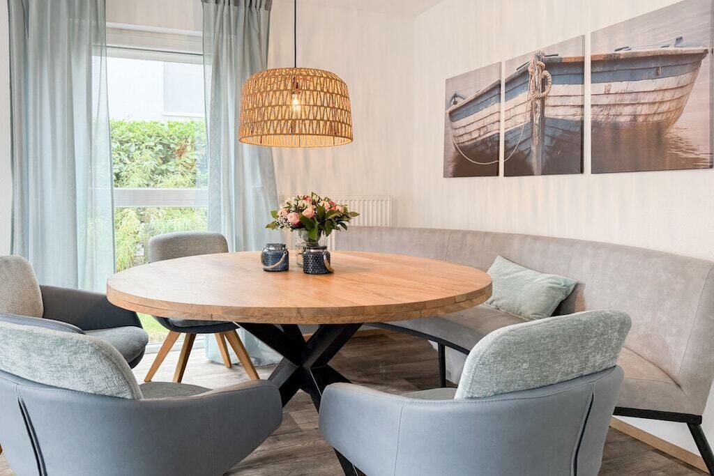 Un comedor con una mesa redonda de madera, sillas tapizadas de color gris, una lámpara colgante tejida y un cuadro en la pared de un barco.
