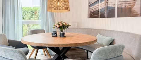 A dining area with a round wooden table, grey upholstered chairs, a woven pendant light, and a wall-mounted painting of a boat.