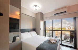 A modern hotel room with a large bed, a bedside table, a mirror, and a window with a city view. - Studio w/ Balcony in Building w/ Pool One0407 (Salvador)