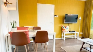 Quarto com mesa de jantar, duas cadeiras, televisão na parede, mesinha branca com luminária e uma porta.