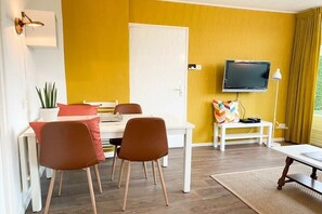 Ein Zimmer mit Esstisch, zwei Stühlen, einem Fernseher an der Wand, einem kleinen weißen Tisch mit Lampe und einer Tür.