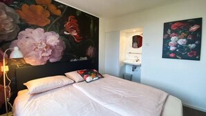 Quarto de hotel com uma cama grande, mesa de cabeceira, espelho fixado na parede, pia e uma pintura de flores.