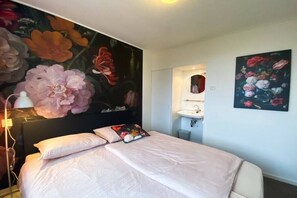 Ein Hotelzimmer mit einem großen Bett, einem Nachttisch, einem wandmontierten Spiegel, einem Waschbecken und einem Blumenbild.