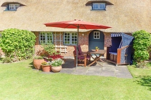 A thatched-roof building with a patio area, a table with chairs, and a red umbrella. - Friesenhaus Sara (S3) (Wenningstedt)