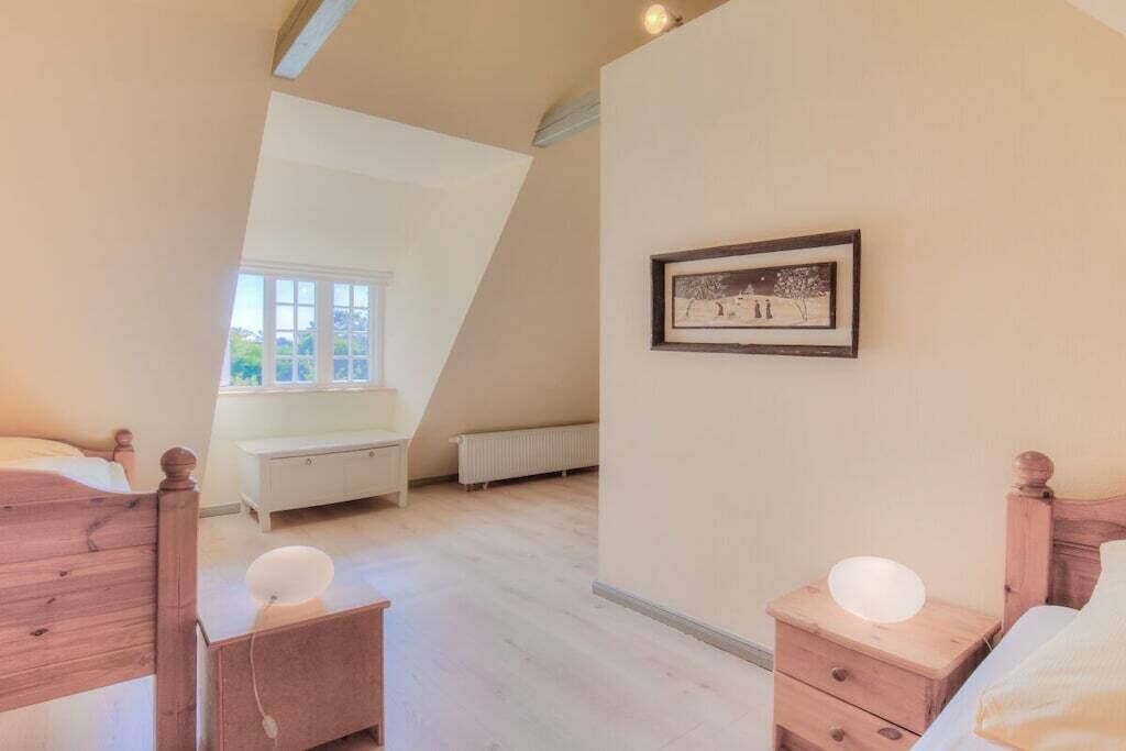 Une chambre avec un plafond en pente, un lit, une table de chevet, une commode, une fenêtre et un tableau encadré au mur.