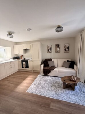 En moderne stue med sofa, sofabord og et køkkenområde med skabe og hårde hvidevarer.
