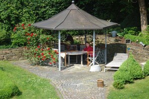 Ein Pavillon mit Tisch und Stühlen, umgeben von einer Steinmauer und viel Grün.