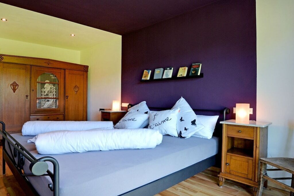 Un lit avec une literie et des oreillers blancs, une armoire en bois, une table de chevet avec une lampe et une étagère murale ornée de tableaux encadrés.