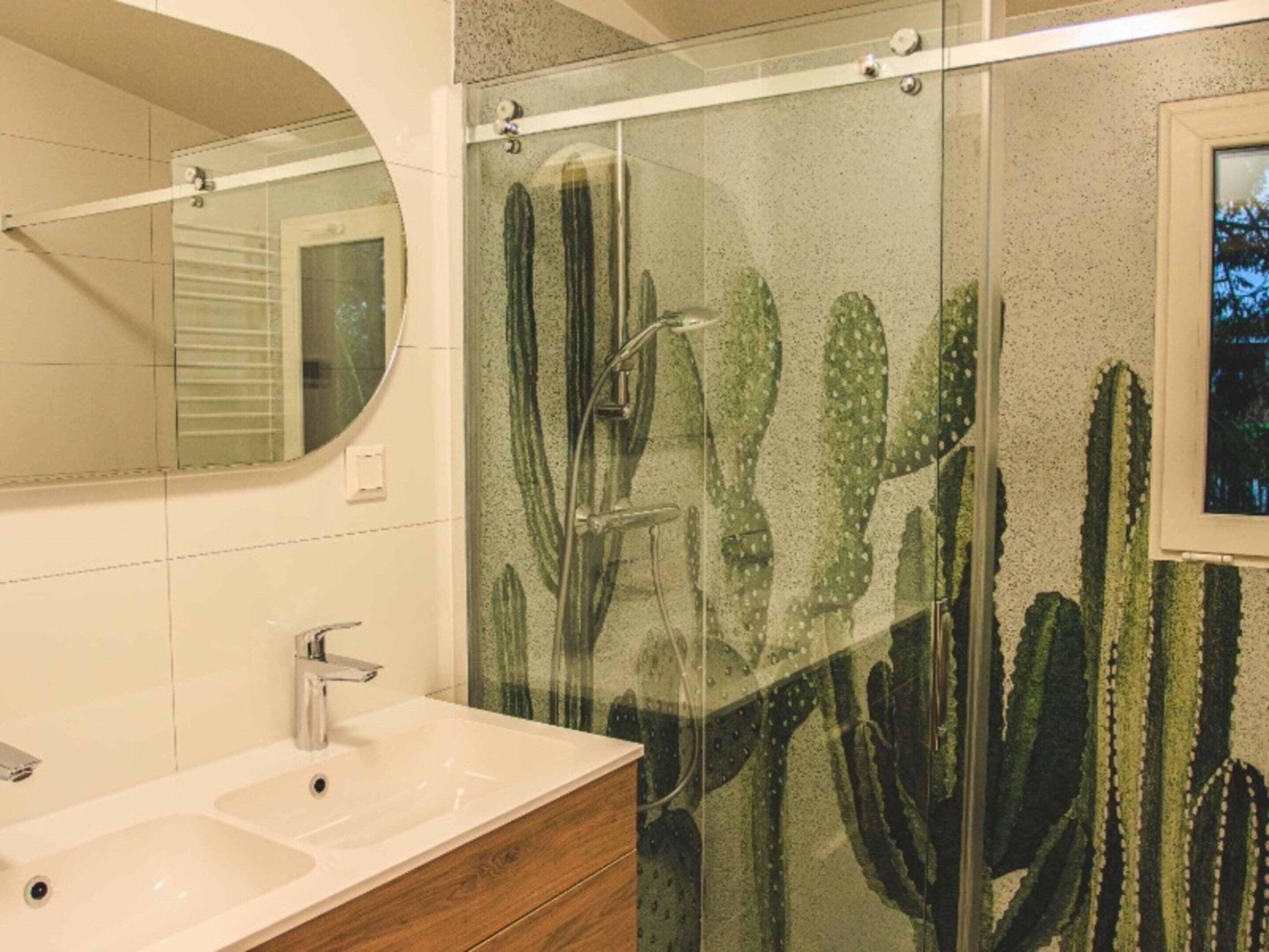 A bathroom with a glass shower enclosure, a sink with a mirror, and a wall with a cactus mural.