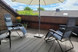 Eine Holzterrasse mit zwei Liegestühlen und einem Tisch, einem Sonnenschirm sowie Blick auf eine Ziegelmauer und Grünpflanzen.