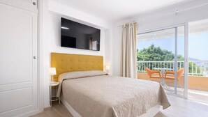 Een hotelkamer met een bed, een aan de muur bevestigde televisie, een balkon met een tafel en stoelen en uitzicht op groen.