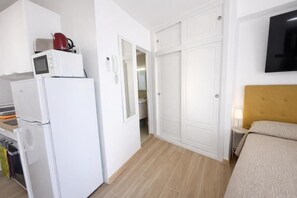 Ein gemütliches Zimmer mit einem Bett, einem kleinen Nachttisch, einem an der Wand befestigten Fernseher und einer Küchenzeile, inklusive Mikrowelle, Toaster und Kühlschrank.