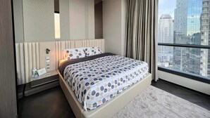 A modern bedroom with a large bed, a nightstand, a mirror, and a view of a cityscape through a large window. - Luxury 3BR Loft with Private Pool & City Views in Makati (Makati City)