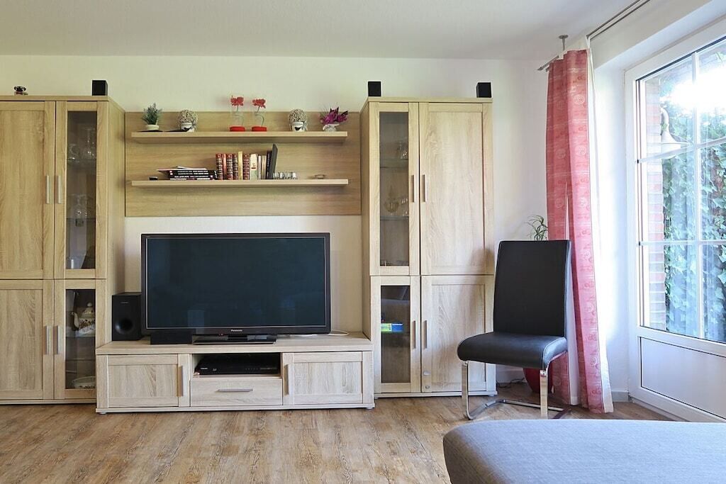 Una sala de estar con un televisor de pantalla plana sobre un mueble de madera, una silla negra y una ventana con cortina roja.