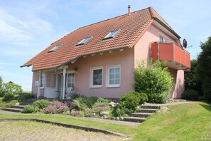 Une maison à deux étages, avec un toit recouvert de tuiles rouges, un balcon agrémenté d’une antenne parabolique et un jardin où poussent diverses plantes et fleurs.